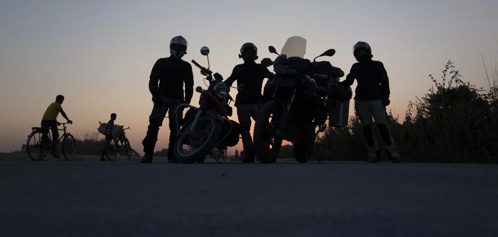 Three riders in silhouette beside their loaded bikes at dusk, with a village road and two cyclists behind them.
