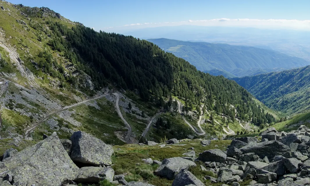 A gravel road switchbacking down a forested mountainside, with a wide valley beyond.