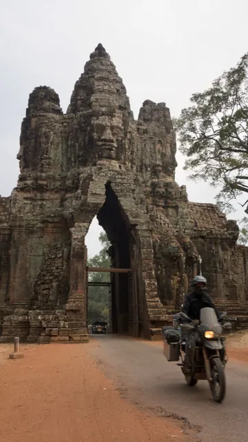 The loaded bike riding out through a carved stone face gate at Angkor onto a red dirt road.