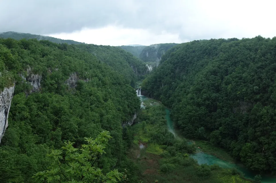 A deep forested gorge with a turquoise river and waterfalls running through it under low cloud.