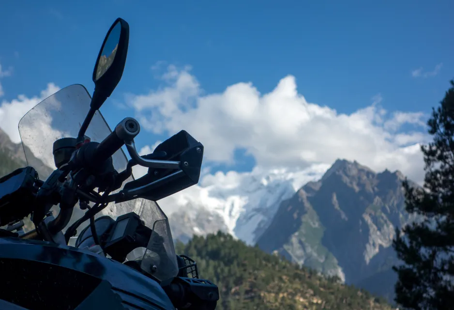 The handlebars and mirror of the loaded bike in the foreground, with snow peaks and cloud filling the sky behind.