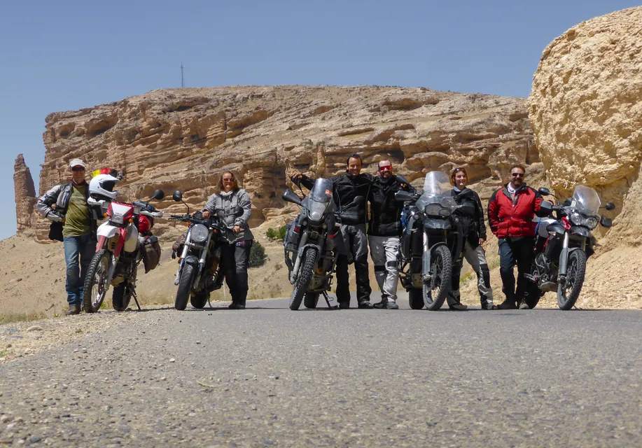 Six riders and their loaded bikes lined up across a desert road below eroded rock cliffs.