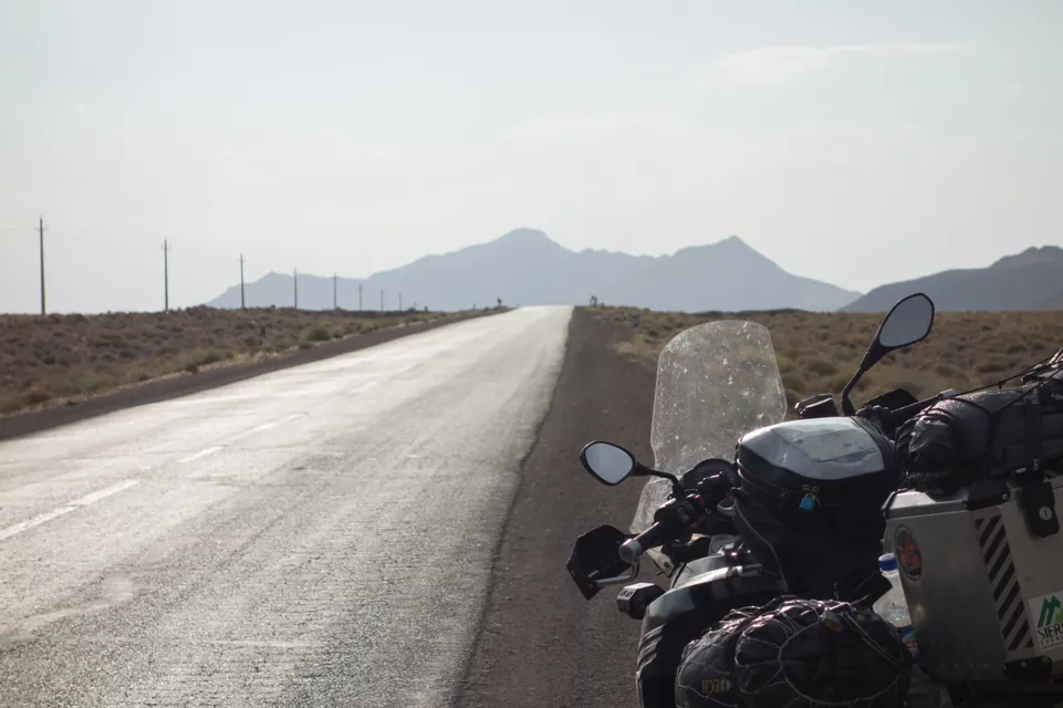 An empty road running straight across the desert toward bare mountains, the loaded bike at the edge of the frame.