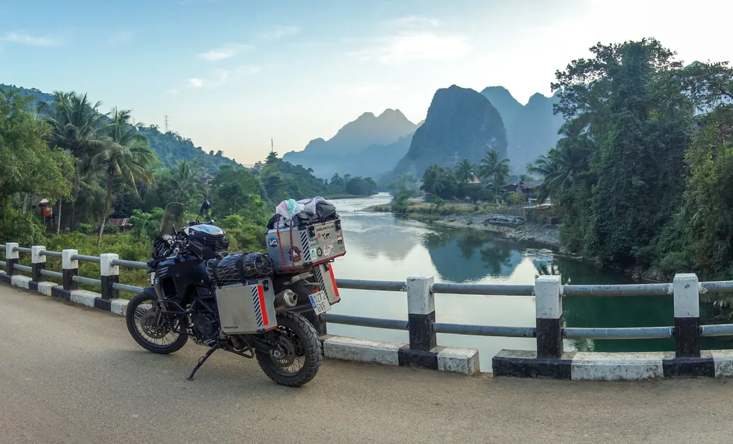 The loaded bike parked on a bridge over a still river, with limestone karst peaks and palms behind it.