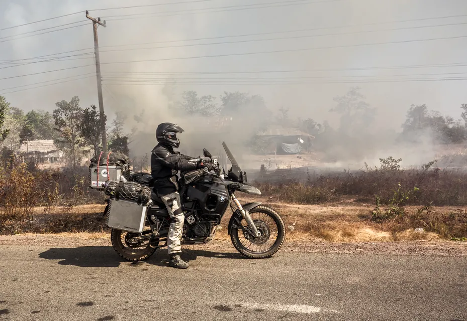 A rider stopped at the roadside beside dry scrub burning off into thick smoke.