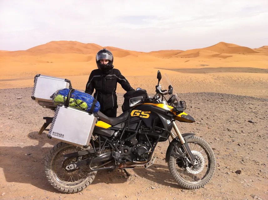 A rider standing beside a loaded yellow BMW GS on hard gravel, with the Sahara dunes rising behind.