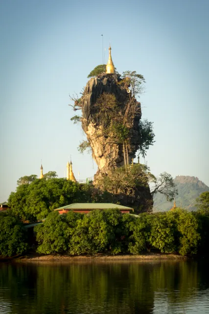 A gold pagoda perched on top of a limestone pinnacle above a lake, with smaller stupas at its foot.
