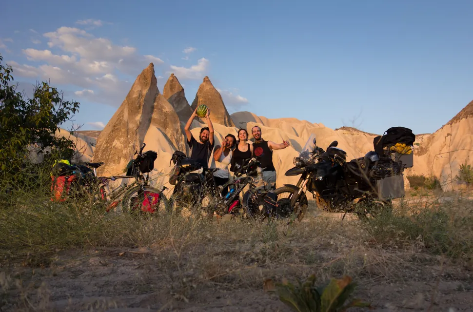 Four travellers with their arms up beside loaded motorbikes and bicycles, in front of the fairy chimneys of Cappadocia at sunset.