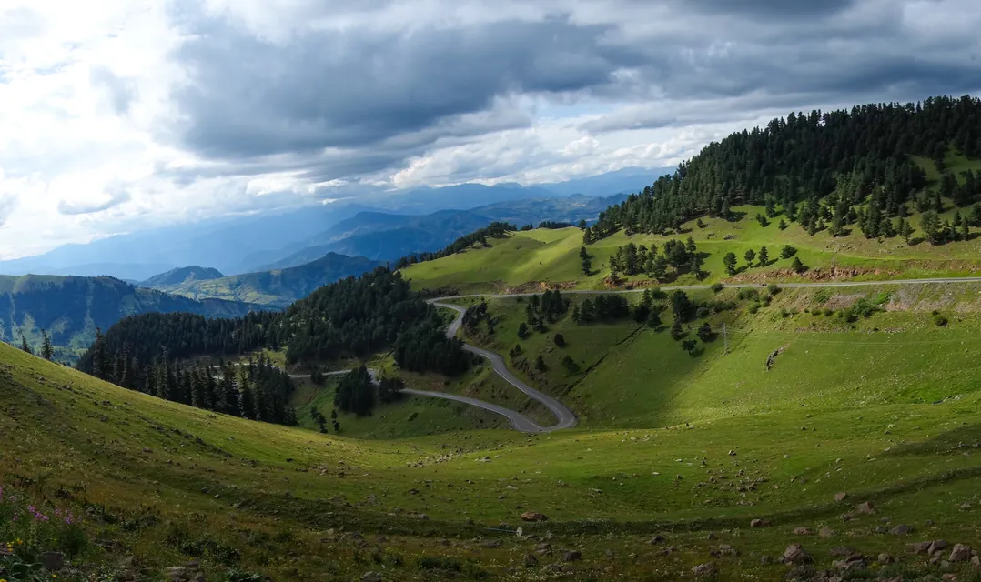 A road switchbacking across high green pasture under heavy cloud, with ranges running away behind.