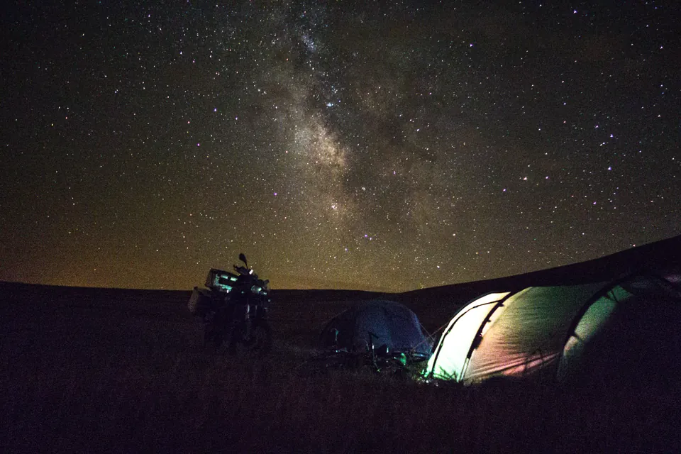 The Milky Way over two tents and the loaded bike, camped on open grassland.